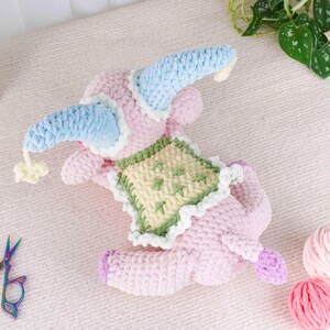 May include: A handmade pink crochet elephant toy with blue and white accents. The elephant wears a blue hat and a green and yellow square on its back. The toy has purple feet and a tail, and a small yellow pom-pom hangs from the hat.