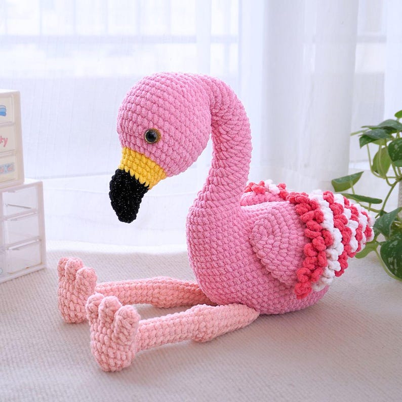 May include: A handmade pink crochet flamingo with a long curved neck, black beak, and yellow accents. The flamingo has a fluffy, textured body with red and white detailing on its back. Its legs are extended, and it has small, detailed feet.