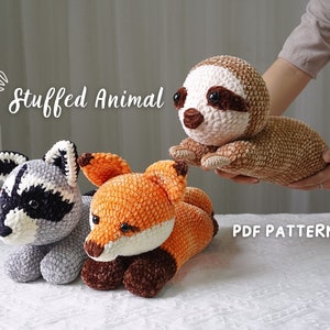 Peut inclure: Trois animaux en peluche crochetés : un raton laveur, un renard et un paresseux. Le raton laveur est gris et noir, le renard est orange et brun, et le paresseux est brun et blanc. Les animaux sont tous couchés sur une surface blanche. Le texte "Stuffed Animal" et "PDF PATTERN" est visible sur l’image.