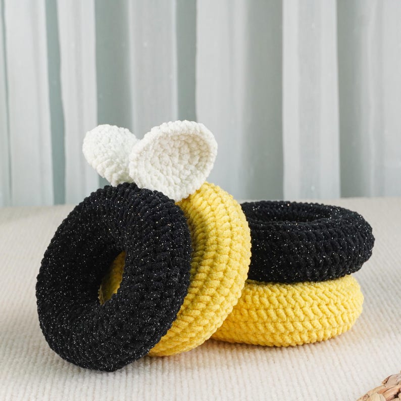 May include: A collection of handmade hair scrunchies in various colors. The scrunchies are black, yellow, and white, with a textured, knitted appearance. Some scrunchies have a glittery sheen. The white scrunchie has a bow-like design.
