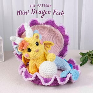 May include: A crocheted yellow Mini Dragon Fish toy with a white horn, wings, and a blue tail, nestled inside a purple and pink crocheted seashell. The text "PDF PATTERN Mini Dragon Fish" is at the top.