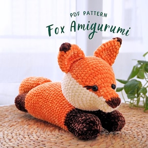 May include: A crocheted orange fox amigurumi toy with brown accents. The toy is lying on a woven straw mat. The text "PDF PATTERN Fox Amigurumi" is displayed above the toy.