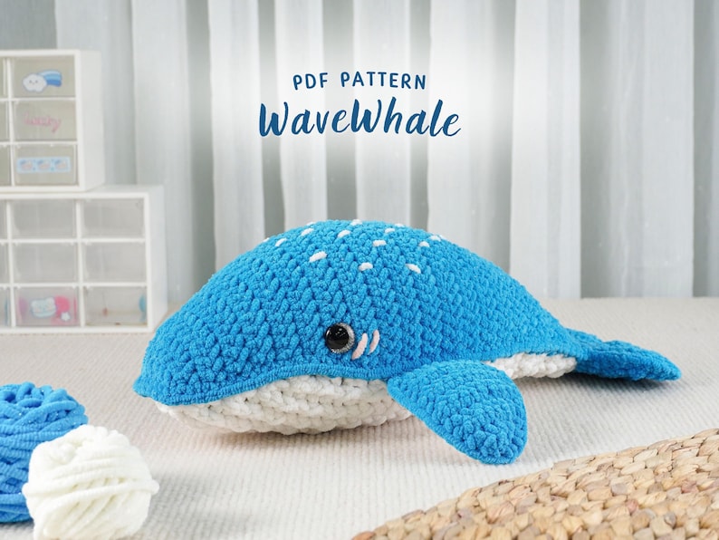 May include: A handmade blue and white crocheted whale plushie with a smiling face and small white dots on its back. The text "PDF PATTERN WaveWhale" is displayed above the whale. Two balls of yarn are in the foreground.
