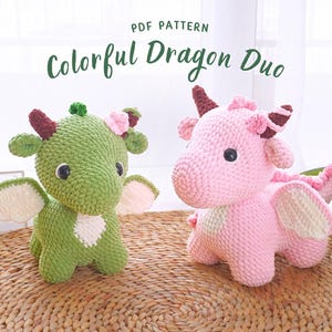 以下が含まれることがあります： 緑とピンクの2匹の編みぐるみドラゴン。白い羽根と茶色の角がついています。籐の敷物の上に座っています。