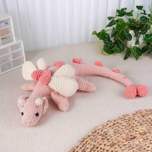 May include: A pink crochet dragon toy with cream wings and heart details. The dragon has small horns and a long tail. The toy is on a light surface, with a plant and storage containers in the background. A charming handmade item.