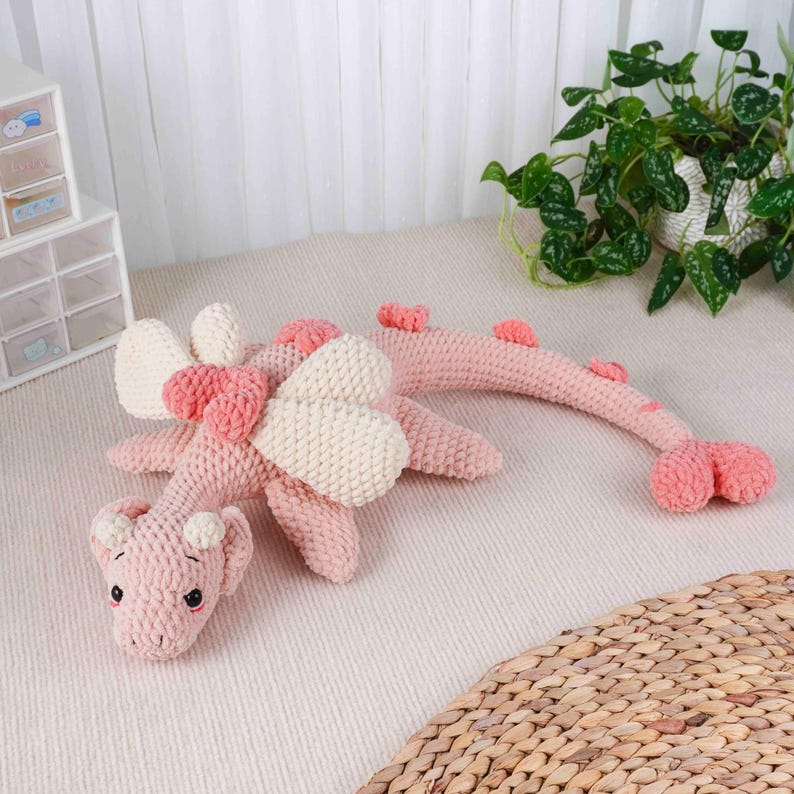 May include: A pink crochet dragon toy with cream wings and heart details. The dragon has small horns and a long tail. The toy is on a light surface, with a plant and storage containers in the background. A charming handmade item.