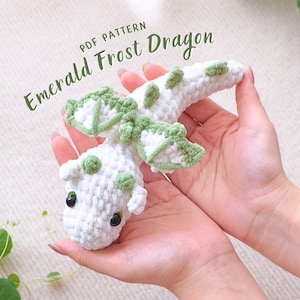 Może przedstawiać: Biały, szydełkowy smok z zielonymi skrzydłami i czarnymi oczami. Smok jest trzymany w dłoniach osoby. Tekst "PDF PATTERN Emerald Frost Dragon" jest widoczny w lewym górnym rogu obrazu.