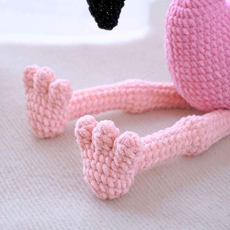 May include: A handmade pink crochet flamingo toy with a black head. The flamingo's legs and feet are a lighter shade of pink. The toy is made of soft, textured yarn.