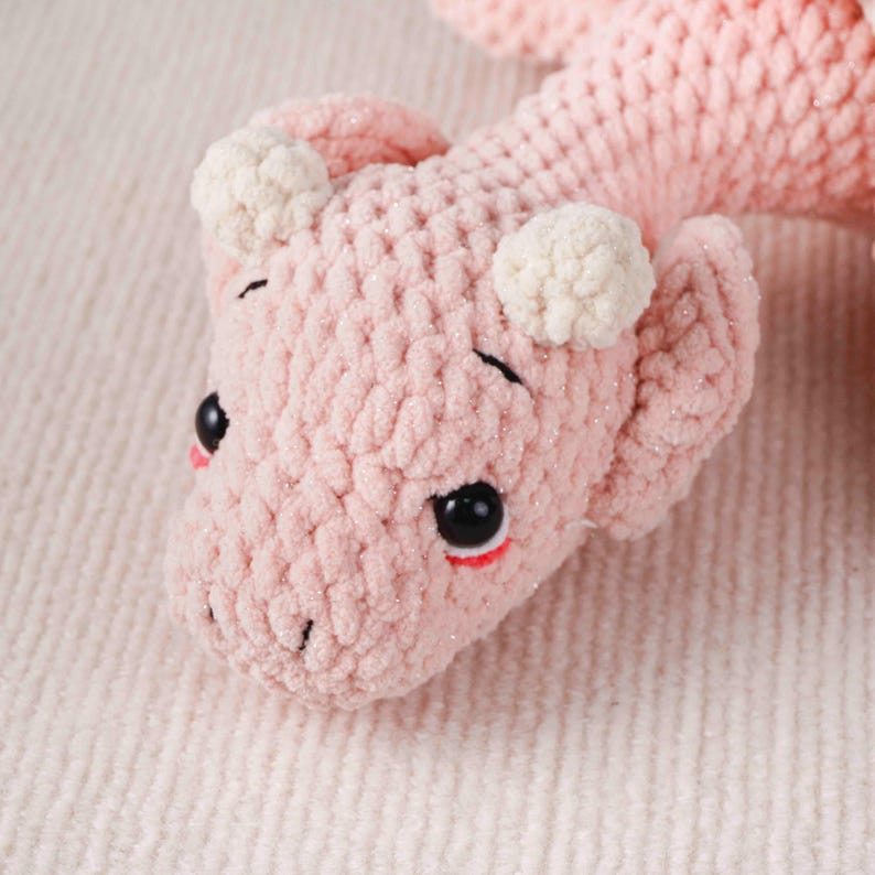 May include: A close-up of a handmade pink crocheted dragon toy. The dragon features black eyes, red accents, and white pom-pom details. The soft, textured yarn gives the toy a cuddly appearance. A charming, handcrafted item.