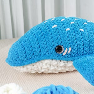 May include: A handmade, blue crochet whale plushie with white spots and a white belly. The whale has a black eye and small white accents. The plushie is made of soft, textured yarn, perfect for a nursery or as a decorative item.