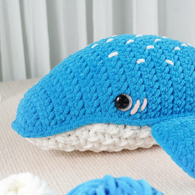 May include: A handmade, blue crochet whale plushie with white spots and a white belly. The whale has a black eye and small white accents. The plushie is made of soft, textured yarn, perfect for a nursery or as a decorative item.
