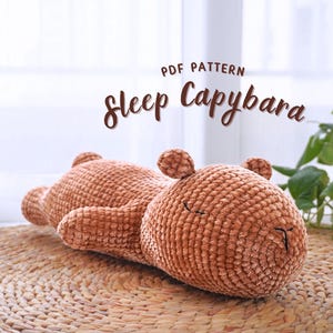Op de afbeelding: Een gehaakt bruin capybara knuffeldier dat op een geweven bruine mat ligt. Het speelgoed heeft een slaperige uitdrukking en ligt op zijn zij. De tekst "PDF PATTERN Sleep Capybara" staat boven het speelgoed.