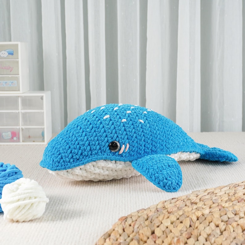 May include: A handmade blue crochet whale plush toy with white accents. The whale features white spots on its back, a white belly, and small black eyes. The toy is made of soft yarn and is designed for children.