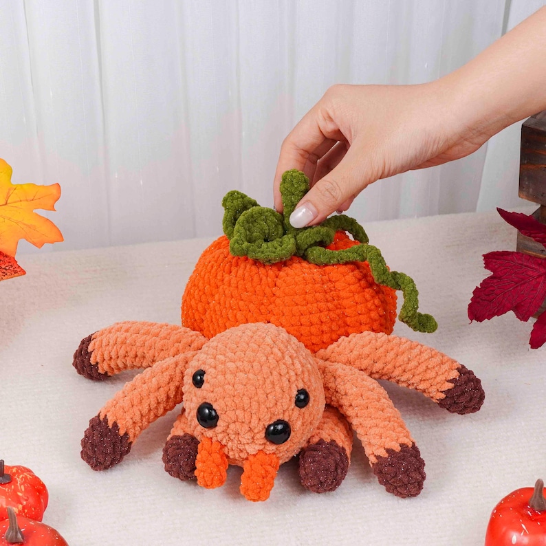 May include: A handmade crochet spider with an orange body and brown-tipped legs sits in front of an orange crochet pumpkin with green vines. The spider has black eyes and an orange mouth. The items are likely for Halloween.