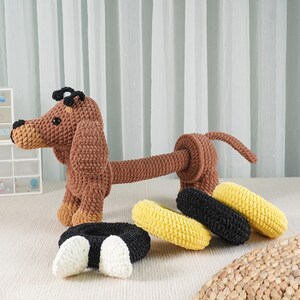 May include: A crocheted dachshund dog in brown, tan, and black, with a long body and floppy ears. Several black, yellow, and black and white crocheted hair scrunchies are arranged near the dog. The dog is a handmade item.