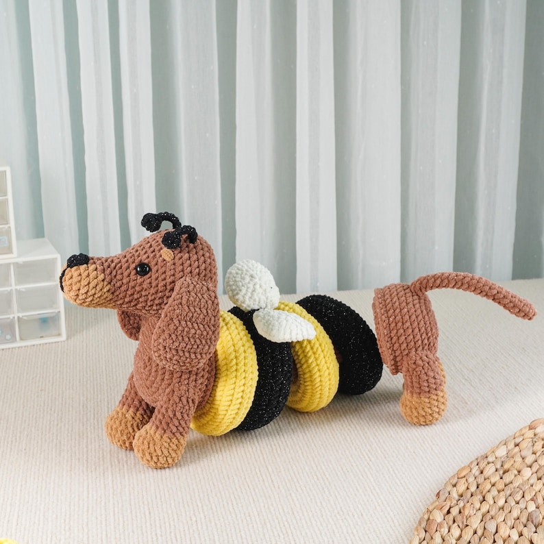 May include: A handmade crochet toy combining a dachshund dog and a bee. The dog's head and legs are brown, with a tan snout. The bee's body is black and yellow, with white wings. The tail is detached and brown.