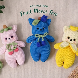 May include: Three crocheted stuffed animals shaped like cats, each with a different fruit theme. The first cat is pink with a strawberry on its head, the second cat is blue with blueberries on its head, and the third cat is yellow with a banana on its head. The text "Fruit Meow Trio" is written above the cats.