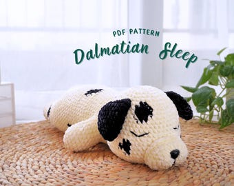 May include: PDF pattern for crocheting a sleeping Dalmatian dog plush toy. The toy is white with black spots and has closed eyes. The toy is lying on a woven mat.