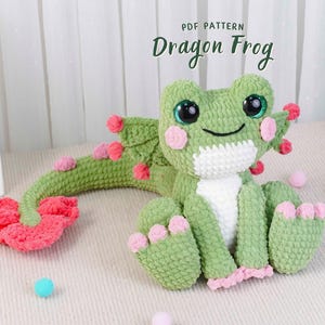 May include: A handmade green crochet Dragon Frog with pink accents. The plush toy has large, expressive eyes, pink cheeks, and a long tail with pink and red details. The text "PDF PATTERN Dragon Frog" is visible.