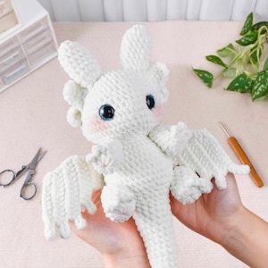 May include: A handmade white crochet dragon with large blue eyes and pink cheeks. The dragon has wings and is being held up by a person. The dragon is a plush toy.