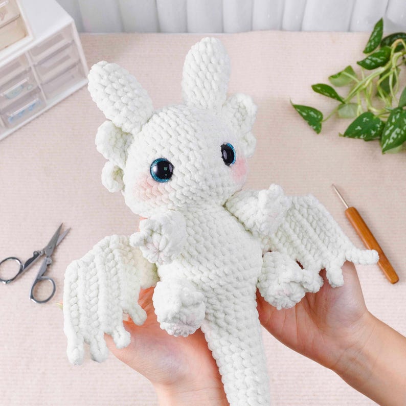 May include: A handmade white crochet dragon with large blue eyes and pink cheeks. The dragon has wings and is being held up by a person. The dragon is a plush toy.