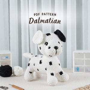 Puede incluir: Un perro dálmata hecho a mano con crochet con manchas negras, orejas negras y nariz negra. La imagen incluye el texto "PDF PATTERN Dalmatian". El perro es blanco con manchas negras y está sentado sobre una superficie blanca.