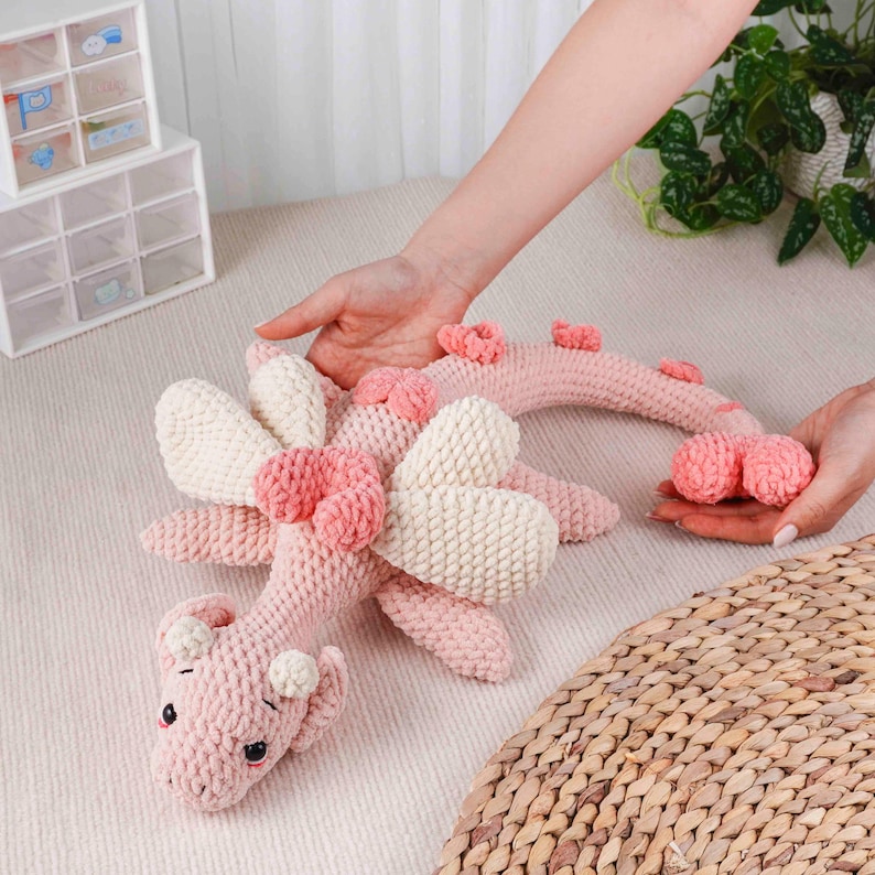 May include: A pink and cream crocheted dragon toy with large wings and decorative accents. The dragon has a long, curved tail. The toy is made of soft yarn and has a whimsical design. The dragon is being held by two hands.