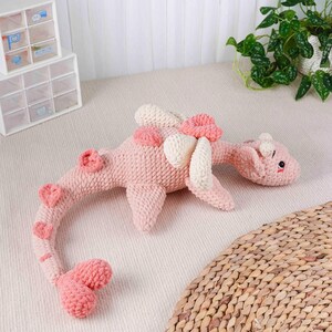 May include: A pink crochet dinosaur toy with cream-coloured details. The toy has a long neck and tail, with heart-shaped features along the back and tail. The dinosaur is lying on a light-coloured surface.