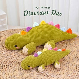 May include: Two green crocheted dinosaur plushies with colorful spines are displayed on a woven mat. The larger dinosaur is positioned behind the smaller one. The text "PDF PATTERN Dinosaur Duo" is at the top of the image.