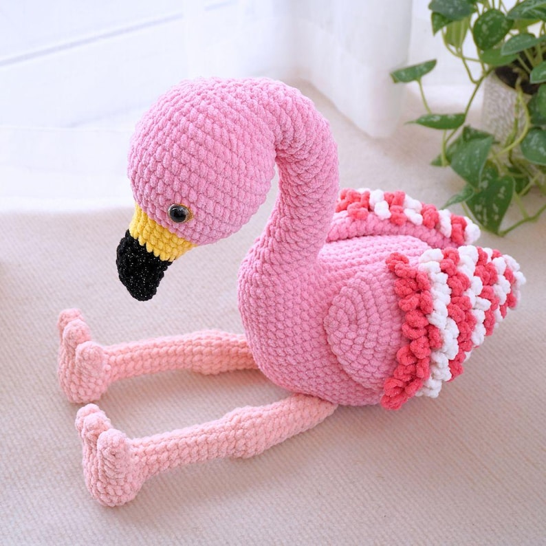 May include: A handmade pink flamingo plush toy with a curved neck and outstretched legs. The flamingo has a black beak, yellow accents, and red and white striped wings. The plush is made of soft, textured yarn.