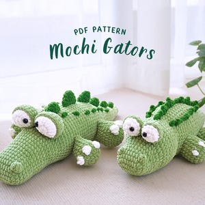 Könnte beinhalten: Zwei gehäkelte Alligator-Plüschtiere, eines grün und eines hellgrün, mit weißen Augen und einer strukturierten Oberfläche. Die Plüschtiere liegen auf einer weißen Oberfläche. Der Text "PDF PATTERN Mochi Gators" wird über den Plüschtieren angezeigt.