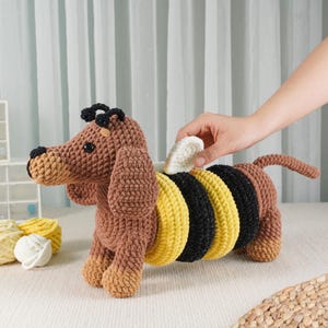 May include: A brown and yellow crocheted dachshund dog toy with black accents. The dog has a bee-like design on its back, with yellow and black stripes. The toy is handmade and has a soft, textured appearance.