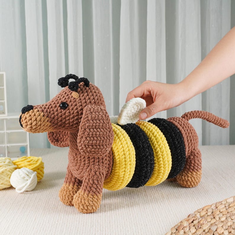May include: A brown and yellow crocheted dachshund dog toy with black accents. The dog has a bee-like design on its back, with yellow and black stripes. The toy is handmade and has a soft, textured appearance.