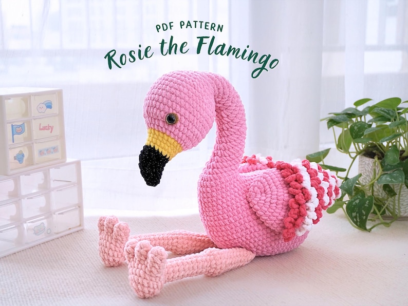May include: A handmade pink crochet flamingo with a black beak, yellow accents, and a white and red ruffled design. The text "PDF PATTERN" and "Rosie the Flamingo" are visible. The flamingo is sitting on a white surface.