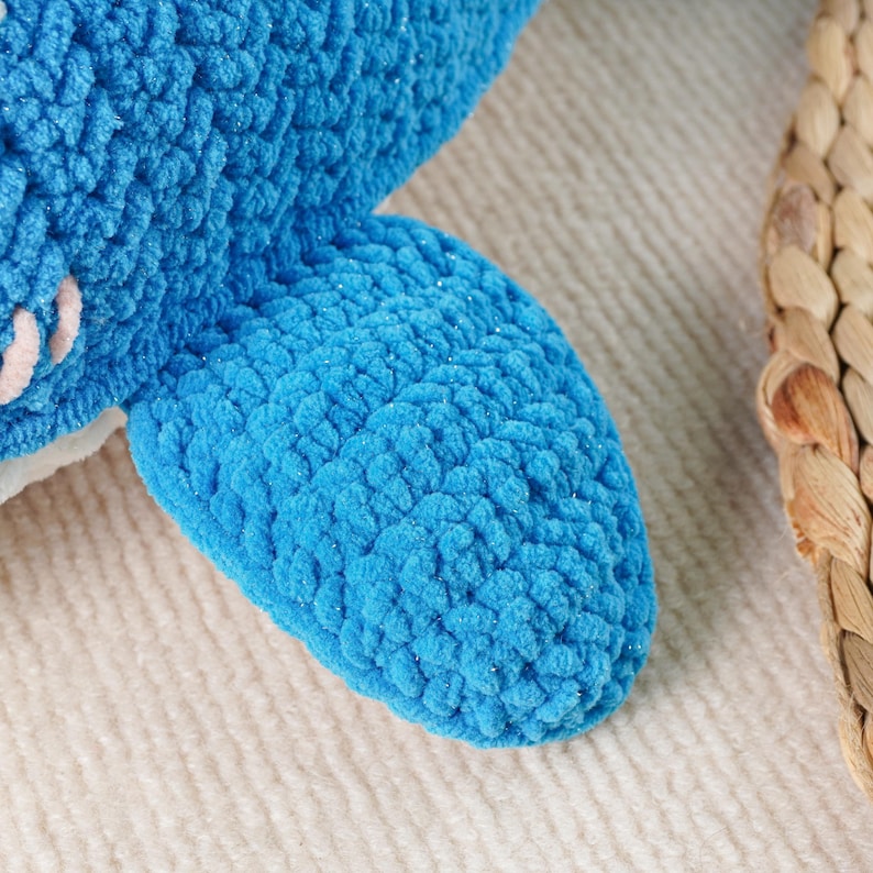 May include: Close-up of a plush blue shark fin made of textured yarn. The fin is a vibrant shade of blue with subtle glitter accents. The background is a soft, off-white knitted fabric, with a portion of a woven basket visible on the right.