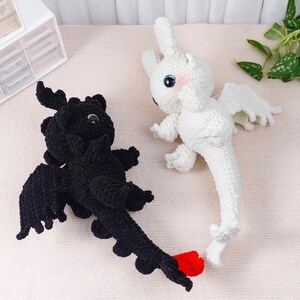 May include: Two crocheted dragon toys. One is black with red accents and the other is white. Both have large eyes, wings, and long tails. The black dragon has a red tail tip. The white dragon has pink cheeks.