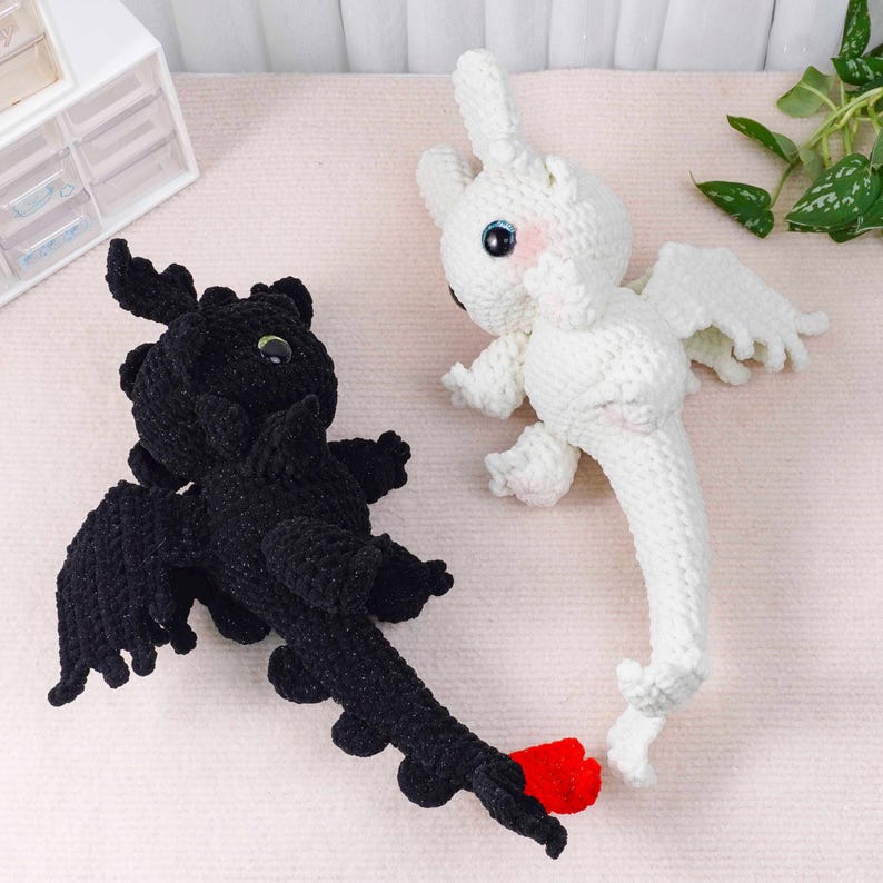 May include: Two crocheted dragon toys. One is black with red accents and the other is white. Both have large eyes, wings, and long tails. The black dragon has a red tail tip. The white dragon has pink cheeks.
