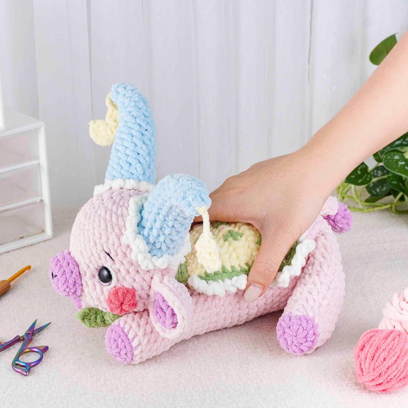 May include: A handmade, pink crochet pig with blue and yellow accents. The pig has large ears, a curly tail, and is lying down. The pig's belly is open, revealing a yellow and green interior. A hand is reaching into the pig's belly.