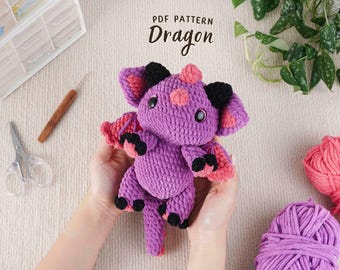 May include: A handmade purple crochet dragon with black and pink accents. The dragon has large ears, wings, and a tail. The image also shows yarn, scissors, and a crochet hook, suggesting a craft project. Text on the image reads "PDF PATTERN Dragon."