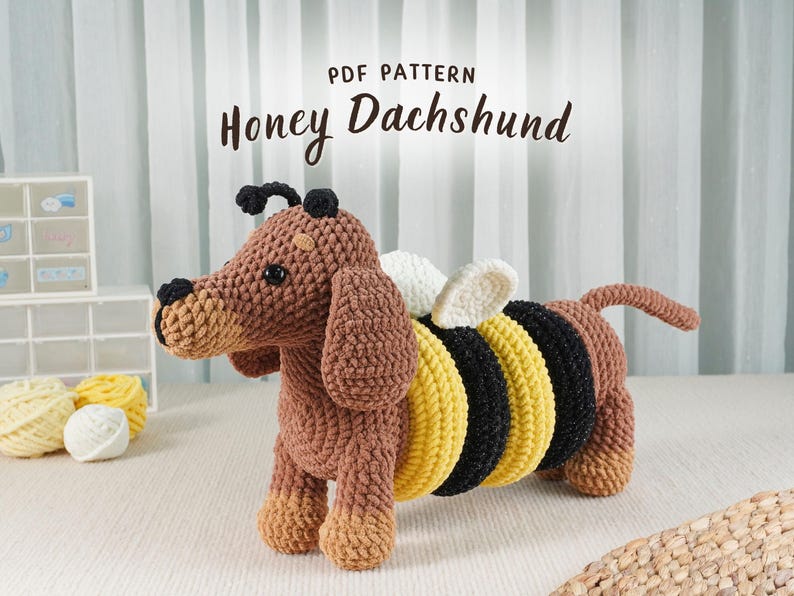 May include: A crocheted dachshund dog with bee-themed accents. The dog is brown with a tan snout and paws, and it has a black and yellow striped body with white wings. The text "PDF PATTERN Honey Dachshund" is at the top.