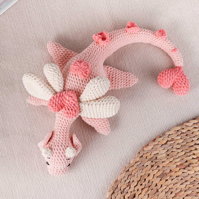 May include: A handmade pink crochet dragon toy with white and pink details. The dragon has a flower design on its chest and wings, a curved neck, and a heart-shaped tail. The toy is made of soft yarn.