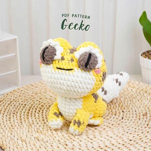 Può includere: Un geco all'uncinetto in giallo, bianco e marrone. Il rettile fatto a mano ha grandi occhi rotondi, una bocca sorridente e una coda a motivi. Il testo "PDF PATTERN Gecko" è sopra il geco.