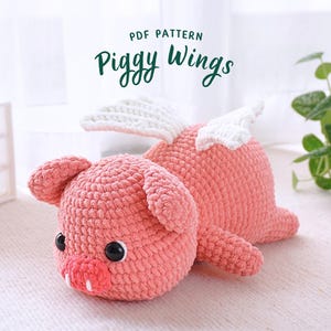 Könnte beinhalten: Ein Häkelmuster für ein rosa Schwein mit weißen Flügeln. Das Muster heißt "Piggy Wings" und ist als PDF-Download erhältlich.
