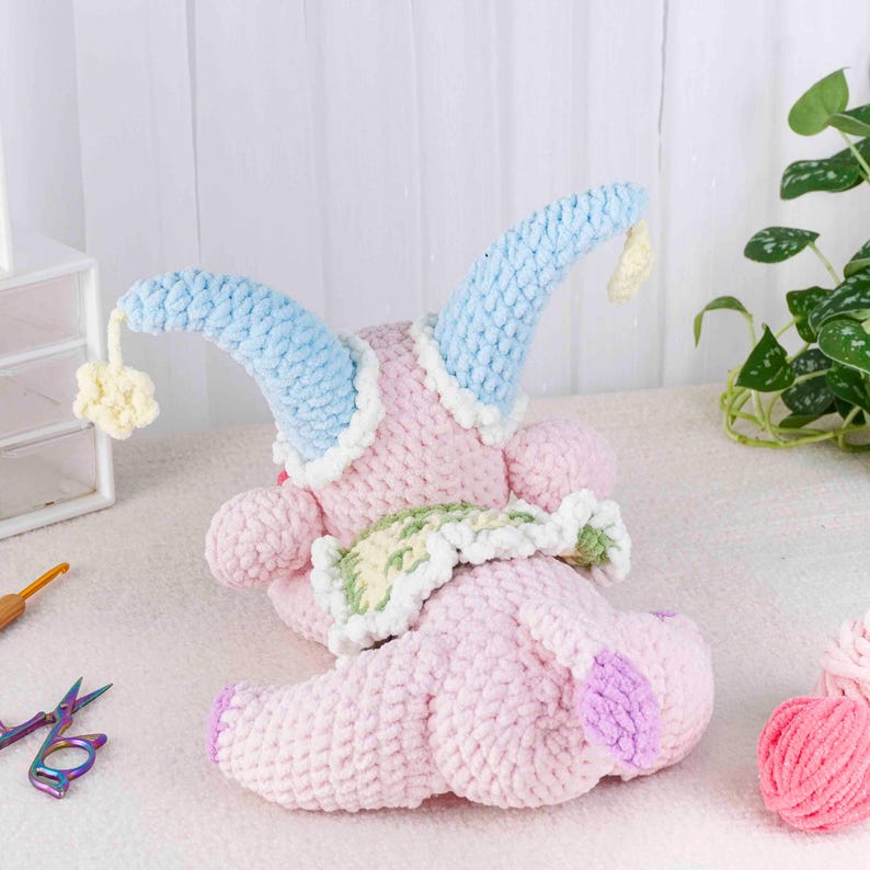 May include: A handmade, plush toy in soft pink, with light blue horns trimmed in white. The toy has a green and white collar and a purple foot. A small yellow pom-pom hangs from one horn. The toy is crocheted.