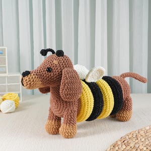 May include: A handmade crochet dachshund dog with bee wings. The dog is brown with tan accents, black eyes, and a black nose. The bee wings are white and cream. The body has yellow and black stripes. A unique, whimsical stuffed animal.