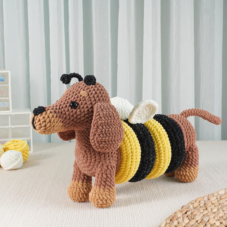May include: A handmade crochet dachshund dog with bee wings. The dog is brown with tan accents, black eyes, and a black nose. The bee wings are white and cream. The body has yellow and black stripes. A unique, whimsical stuffed animal.