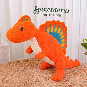 May include: A crocheted Spinosaurus dinosaur toy in bright orange with a cream-colored belly and a large, fan-shaped dorsal fin featuring blue and yellow accents. The text Spinosaurus PDF PATTERN is visible above the toy in dark green lettering.