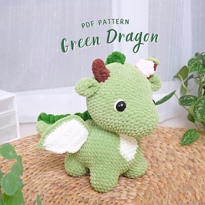 以下が含まれることがあります： 白い羽根とピンクの花が頭についた緑色の編みぐるみドラゴンのおもちゃ。ドラゴンは編まれたマットの上に座っています。PDF PATTERN Green Dragon.