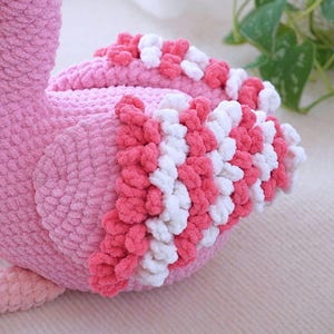 May include: A plush pink flamingo toy with a textured, soft appearance. The flamingo features a detailed design with white and pink accents on its wings. The toy is designed to be soft and cuddly.