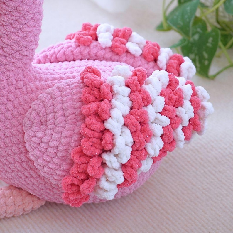 May include: A plush pink flamingo toy with a textured, soft appearance. The flamingo features a detailed design with white and pink accents on its wings. The toy is designed to be soft and cuddly.